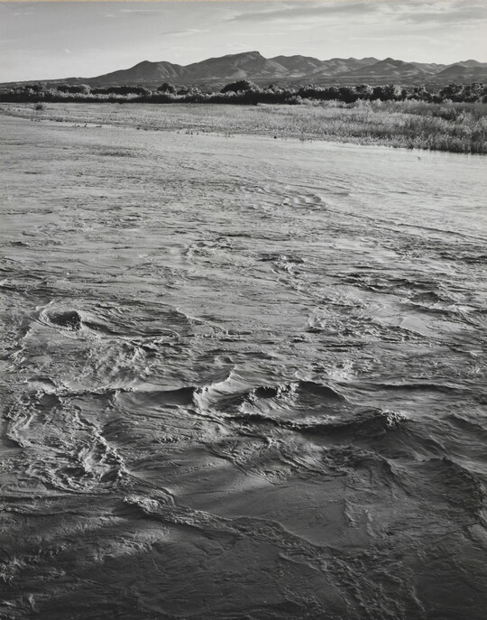 A black-and-white photograph of a choppy river and then grassy plains and mountains beyond the shore.
