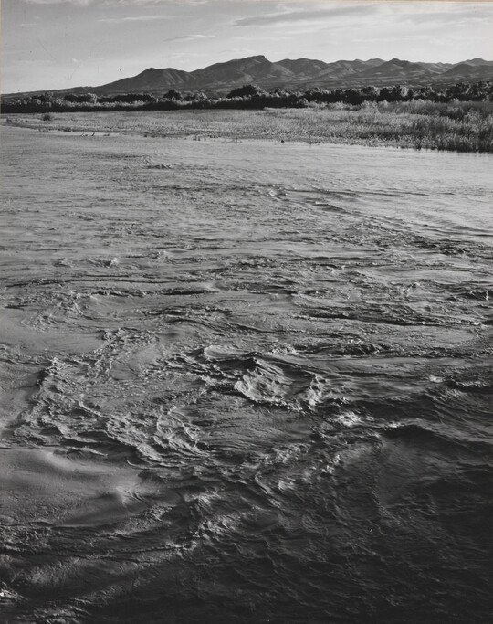 A black-and-white photograph of a choppy river and then grassy plains and mountains beyond the shore.
