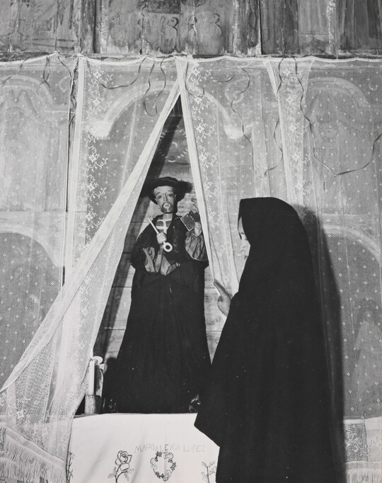 A black-and-white photograph of a woman wearing a black shawl over her head facing a statue of a saint holding a large key.