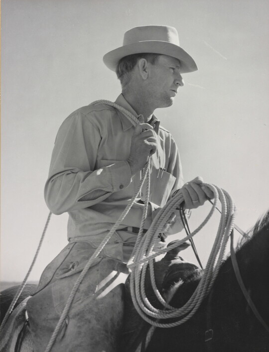 A black-and-white photograph of a White man wearing a brimmed hat holding a lasso astride a dark horse.