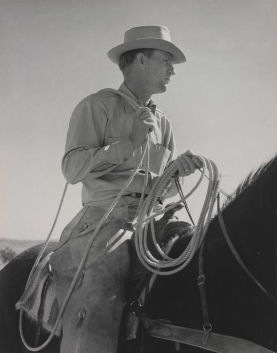 A black-and-white photograph of a White man wearing a brimmed hat holding a lasso astride a dark horse.
