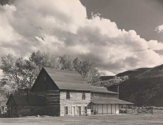 A black-and-white photograph of a three-story log building with mountains and large puffy clouds in the background.