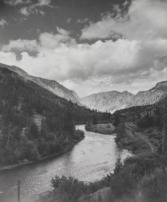 A black-and-white photograph of a river winding through a mountain valley under a cloudy sky.