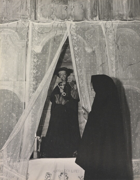 A black-and-white photograph of a woman wearing a black shawl over her head facing a statue of a saint holding a large key.