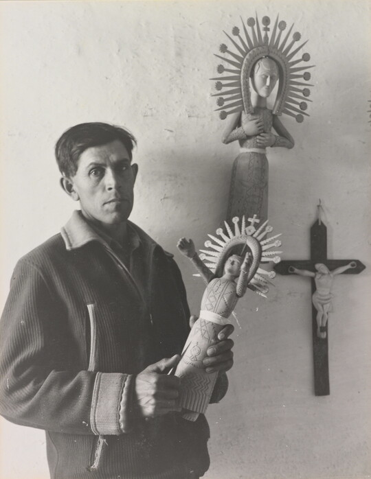 A black-and-white photograph of a man in a winter jacket holding an elaborately carved wooden figure.