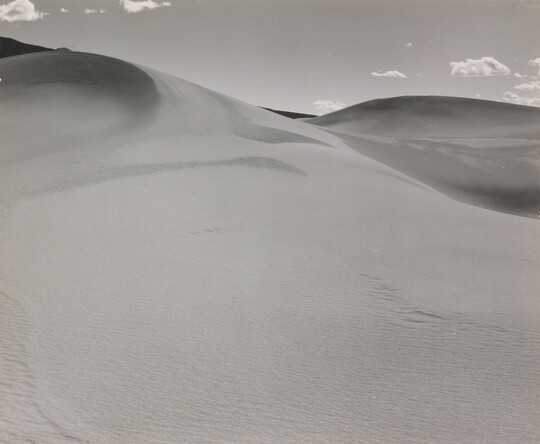 A black-and-white photograph of a rippling sand dune rising up into a sky dotted with a few small clouds.