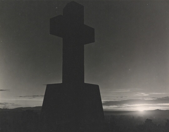 A black-and-white photograph of a stone cross in silhouette and the sun peeking out from the horizon.