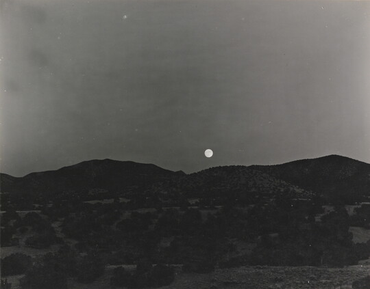 A black-and-white photograph of the moon rising above a mountain landscape.