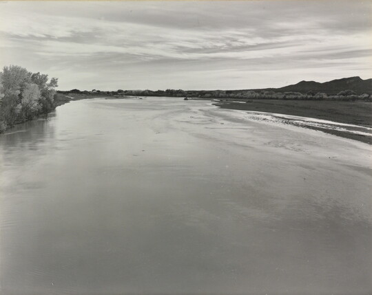 A black-and-white photograph of a calm river with trees on the left shore and mountains in the distance.