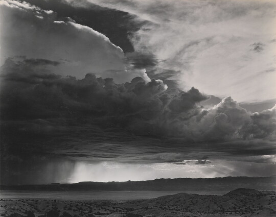 A black-and-white photograph of torrential rain and dark, heavy storm clouds passing into a brightly lit plain.