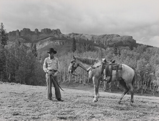 A black-and-white photograph of a White cowboy holding the reins of a horse in front of mountains.