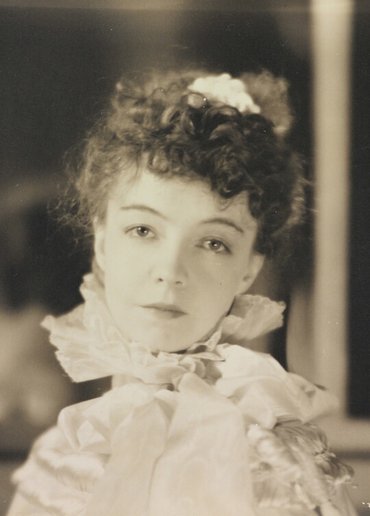 A black-and-white portrait photograph of a White woman with curly hair pulled on top of her head, wearing a top with a ruffled collar.