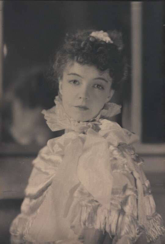 A black-and-white portrait photograph of a White woman with curly hair pulled on top of her head, wearing a top with a ruffled collar.
