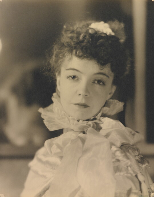 A black-and-white portrait photograph of a White woman with curly hair pulled on top of her head, wearing a top with a ruffled collar.