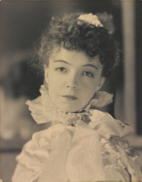 A black-and-white portrait photograph of a White woman with curly hair pulled on top of her head, wearing a top with a ruffled collar.