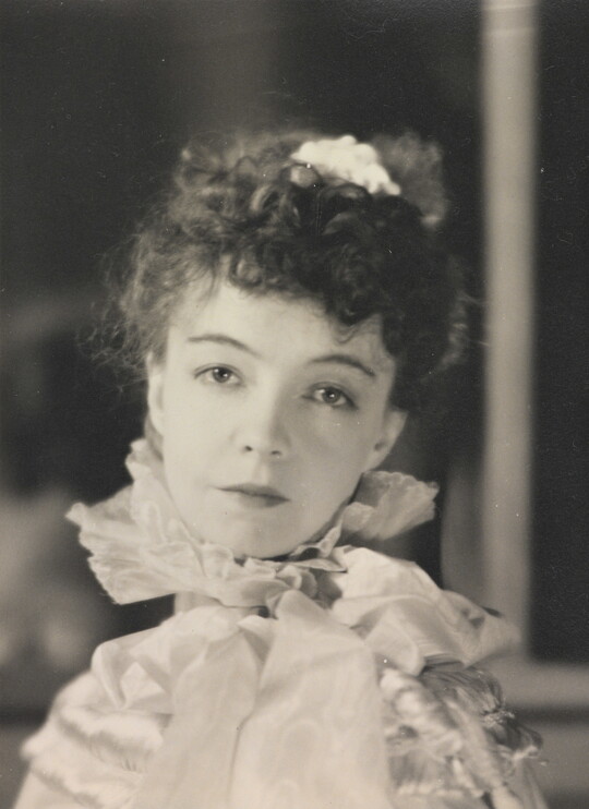 A black-and-white portrait photograph of a White woman with curly hair pulled on top of her head, wearing a top with a ruffled collar.