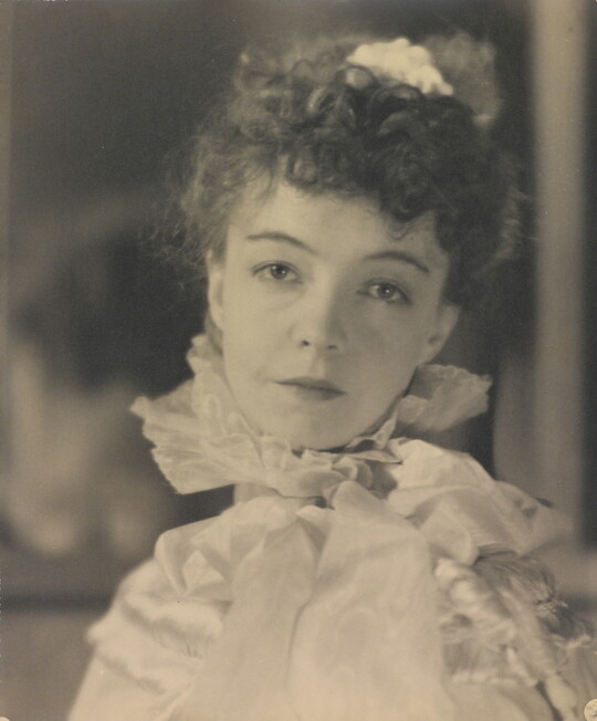 A black-and-white portrait photograph of a White woman with curly hair pulled on top of her head, wearing a top with a ruffled collar.