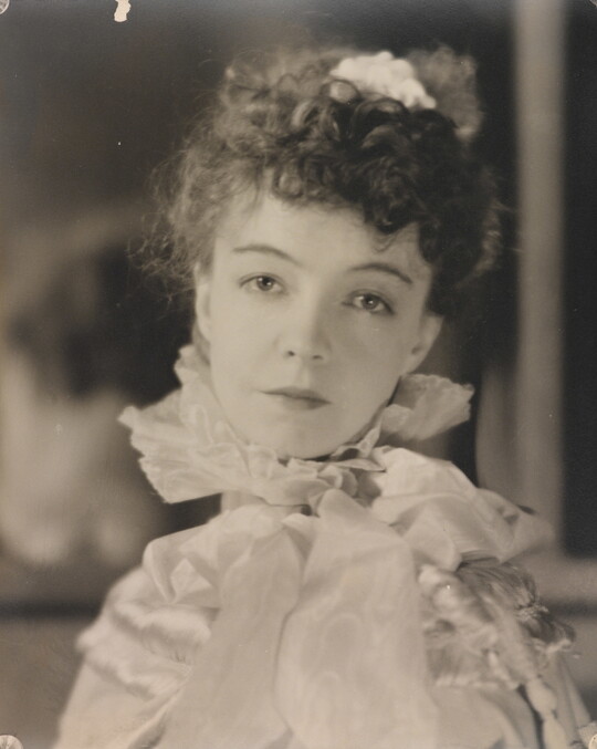 A black-and-white portrait photograph of a White woman with curly hair pulled on top of her head, wearing a top with a ruffled collar.