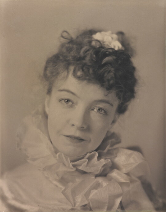 A black-and-white portrait photograph of a White woman with curly hair pulled on top of her head, wearing a top with a ruffled collar.