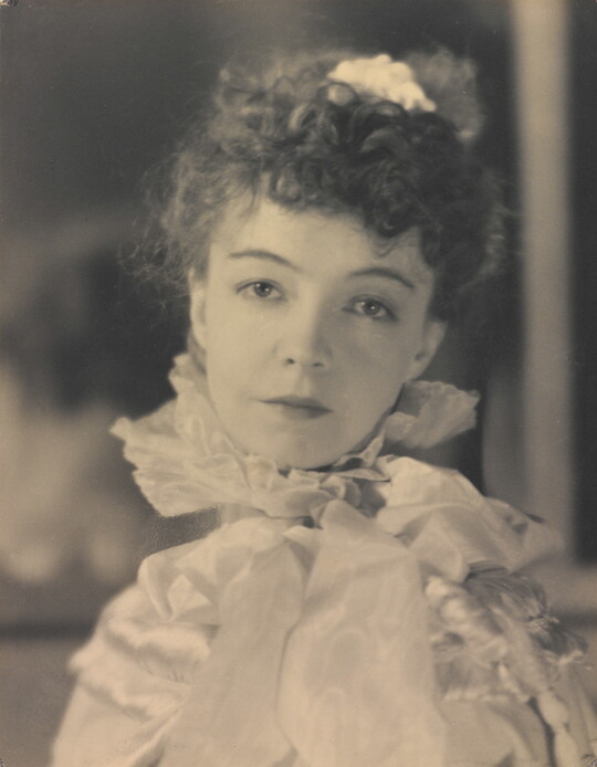 A black-and-white portrait photograph of a White woman with curly hair pulled on top of her head, wearing a top with a ruffled collar.