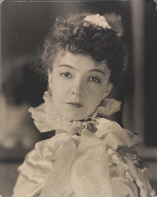 A black-and-white portrait photograph of a White woman with curly hair pulled on top of her head, wearing a top with a ruffled collar.