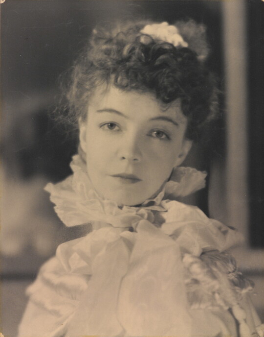 A black-and-white portrait photograph of a White woman with curly hair pulled on top of her head, wearing a top with a ruffled collar.