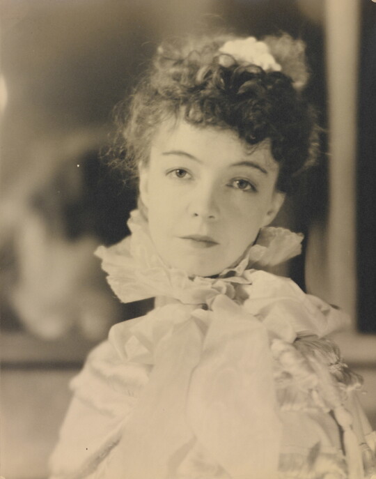 A black-and-white portrait photograph of a White woman with curly hair pulled on top of her head, wearing a top with a ruffled collar.