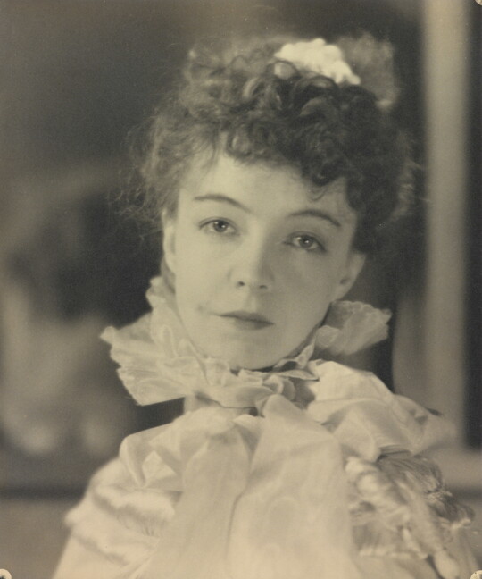 A black-and-white portrait photograph of a White woman with curly hair pulled on top of her head, wearing a top with a ruffled collar.