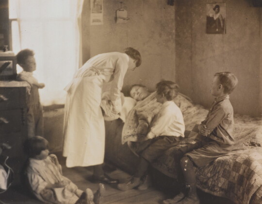A black-and-white photograph of a nurse bending over a person in a bed, two children sitting on the bed, and two children behind the nurse.