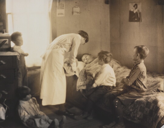 A black-and-white photograph of a nurse bending over a person in a bed, two children sitting on the bed, and two children behind the nurse.