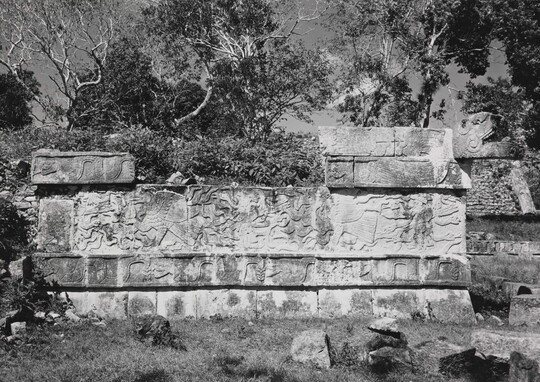 A black-and-white photograph of a crumbling stone wall engraved with designs surrounded by grass and trees.