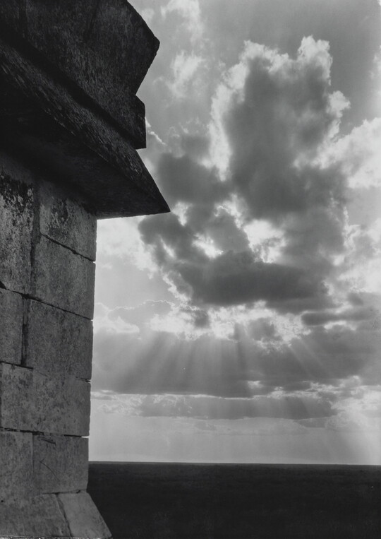 A black-and-white photograph of the sun's rays bursting from clouds and a stark brick architectural feature in the left foreground.
