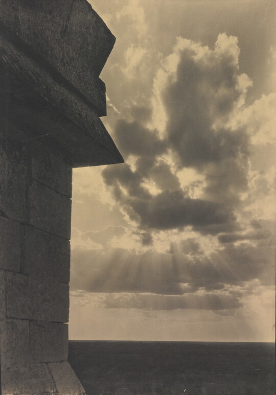 A sepia-toned photograph of the sun's rays bursting from clouds and a stark brick architectural feature in the left foreground.