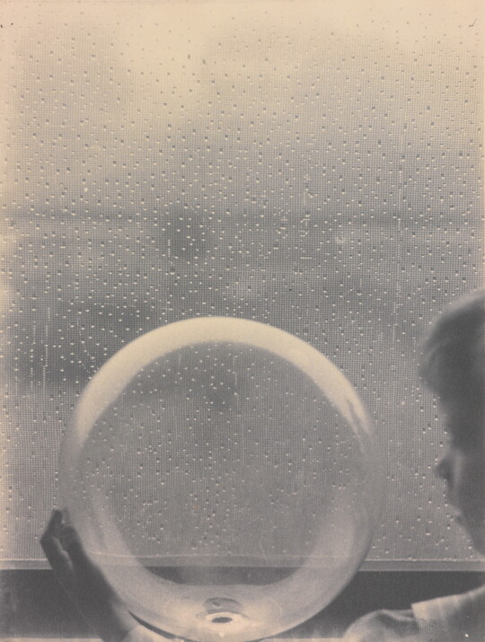 A black-and-white photograph of a White child looking at a clear glass globe in front of a rain-spattered window.