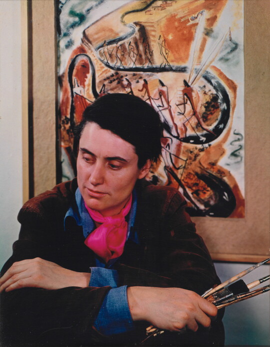 A color portrait photograph of a seated White woman holding paintbrushes in front of an abstract painting.