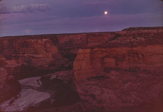 A color photograph of a full moon shining in a purple and blue sky over a steep, red, rocky canyon.
