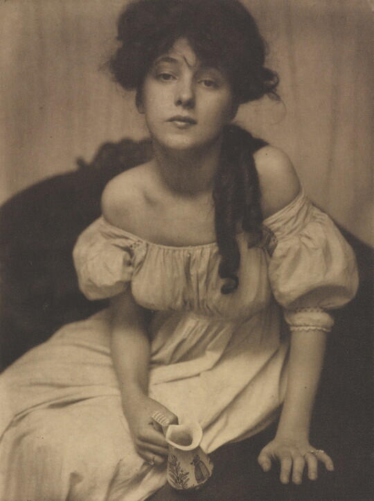 A sepia-toned portrait photograph of a seated young woman dressed in white leaning forward and gazing into the camera.