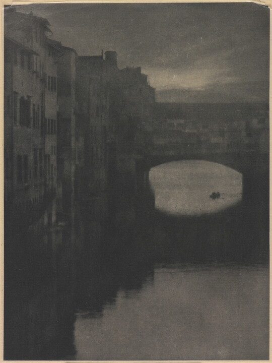 A sepia-toned photograph of a wall of tall buildings lining a river and an enclosed arched bridge stretching over the water.