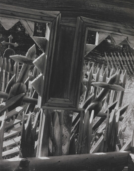 A black-and-white photograph looking through the windows of a wooden structure onto a cemetery with wooden crosses and fence posts.
