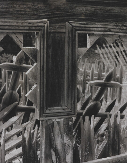A black-and-white photograph looking through the windows of a wooden structure onto a cemetery with wooden crosses and fence posts.