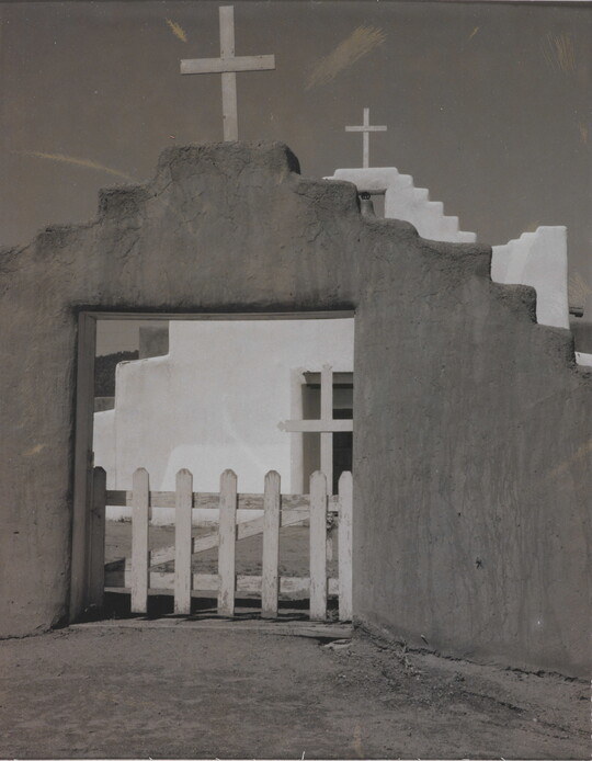 A black-and-white photograph of a white adobe church seen through the entrance of the surrounding adobe wall topped with a wooden cross.