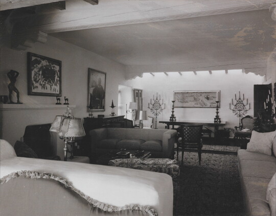 A black-and-white photograph of a formal living room in an adobe building with a fireplace, multiple couches, paintings, and sculptures.
