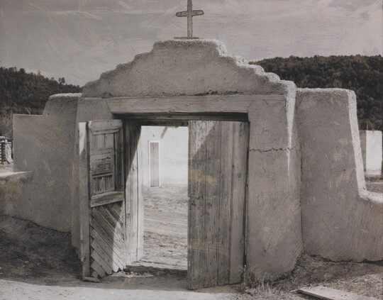 A black-and-white photograph of an adobe wall with a doorway that leads to a courtyard; a cross on the wall above the wooden doors.