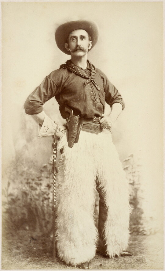 A sepia-toned formal portrait photograph of a cowboy posing in western wear, including white furry chaps, with his hands on his hips.