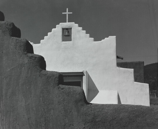 A black-and-white photograph of a terraced adobe bell tower with a cross on top, partially hidden by a terraced adobe wall.