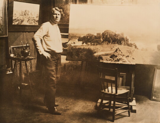 A sepia-toned photograph of a White man posing next to a large canvas that depicts a herd of bison.