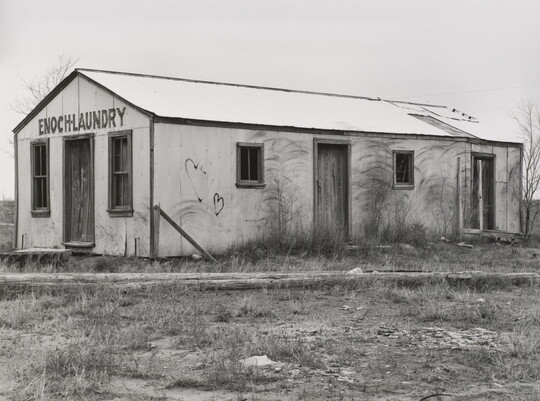 A black-and-white photograph of a one-story building with boarded up windows and graffiti on the walls; lettering above the door reads, "Enoch Laundry."