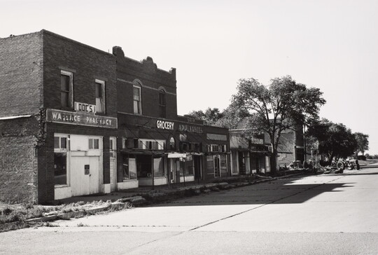 A black-and-white photograph of a paved street lined with brick buildings, some boarded up, casting stark shadows on the ground.