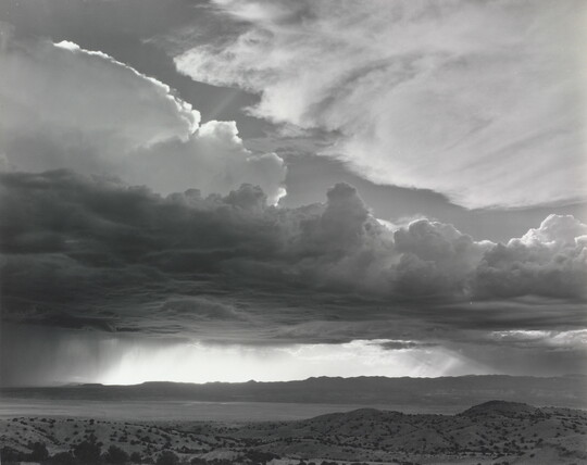 A black-and-white photograph of torrential rain and dark, heavy storm clouds passing into a brightly lit plain.