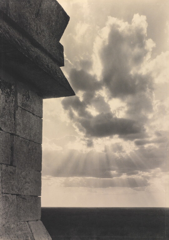 A sepia-toned photograph of the sun's rays bursting from clouds and a stark brick architectural feature in the left foreground.